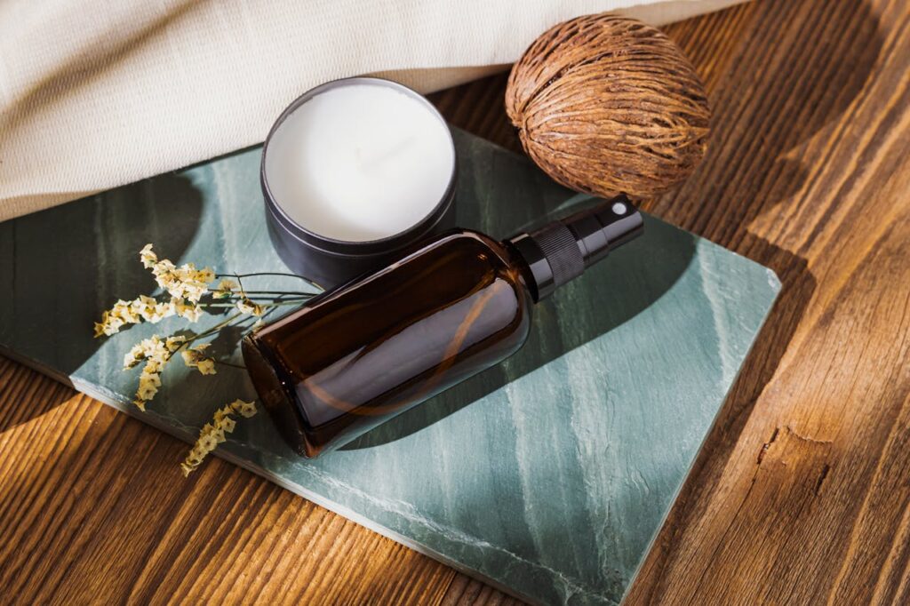 Close-up of a brown glass spray bottle with candle and coconut on green marble setting.