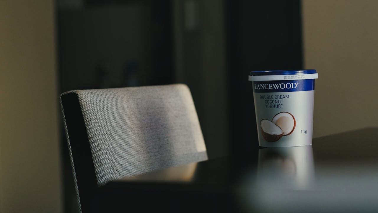 A tub of double cream coconut yoghurt sits on a table next to a chair indoors.
